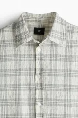 Camisa de algodón con textura arrugada, de color blanco con un patrón de cuadros sutil en gris. Presenta cuello inglés, tapeta lisa con botones blancos y mangas largas con puños ajustables.