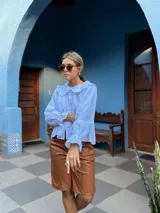 Camisa blanca de corte holgado con cuello bebé con volados, cierre frontal de botones y mangas largas con puños terminados en volados.