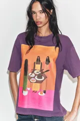 Remera oversize de algodón con efecto lavado en color violeta. Presenta cuello redondo, manga corta y una estampa frontal colorida con una ilustración artística de Manuja Waldia.