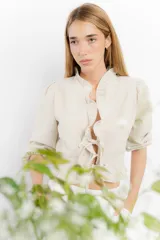 Blusa corta blanca de lino con mangas 3/4 abullonadas, escote en V con volados y cierre con lazos.