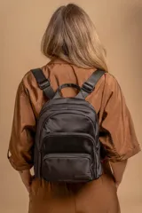 Mochila urbana de diseño funcional en color beige, confeccionada en tela impermeable. Cuenta con un compartimento principal amplio, un bolsillo secundario y un bolsillo frontal, todos con cierre. Incluye asa superior y correas ajustables para los hombros, con detalle de etiqueta de marca en el frente.