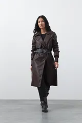 Trench coat de símil cuero color marrón oscuro, con solapas amplias, cierre frontal con botones, cinturón ajustable en la cintura y mangas con tiras ajustables. Presenta un largo por debajo de la rodilla y bolsillos laterales.