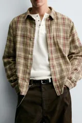 Camisa de corte cropped confeccionada en algodón, con estampado de cuadros en tonos beige, marrón y rojo. Presenta cuello solapa, cierre frontal de botones, manga larga con puño abotonado y detalles de roturas distribuidas por toda la prenda con un bajo irregular.