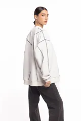 Buzo blanco de manga larga con diseño holgado, cuello redondo y costuras externas visibles en contraste. Cuenta con una etiqueta decorativa en el frente inferior.