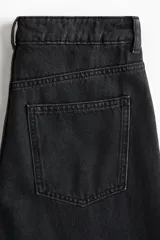 Jean de tiro alto y estilo baggy color negro, confeccionado en denim de algodón rígido, con diseño de cinco bolsillos, cierre de cremallera y botón metálico, y perneras amplias de corte holgado.