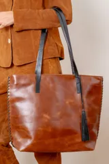Bolso tipo tote confeccionado en cuero vacuno color chocolate, con detalles de costuras decorativas en los laterales, asas largas negras y borla decorativa de cuero.