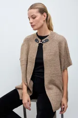 Capa tejida de punto en color beige, con diseño abierto y cierre frontal mediante broche metálico con detalles en cuero sintético negro. Presenta mangas cortas y textura suave.