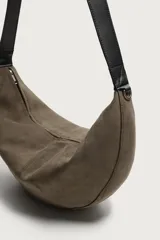 Bolso de hombro tipo hobo con diseño de media luna, confeccionado en gamuza color marrón. Cuenta con un asa larga fija de cuero negro y cierre superior con cremallera.