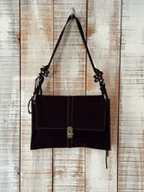 Cartera de hombro color marrón oscuro, confeccionada en gamuza, con diseño rectangular y cierre frontal metálico. Presenta una correa ajustable con detalles decorativos trenzados en los laterales.