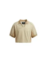 Camiseta crop de manga corta con cuello polo, confeccionada en tela técnica tipo dry fit. Presenta un diseño a rayas verticales finas en color beige, con paneles laterales de malla en tono camel y detalles de parches bordados en el frente.