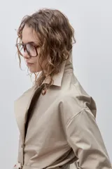 Trench clásico color marrón, de silueta relajada, confeccionado en tejido liviano, con cuello de solapa, doble abotonadura frontal y mangas largas con presillas regulables en los puños.