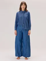 Camisa de denim azul con cuello redondo, volados decorativos en el cuello y a lo largo de la cartera frontal con botones. Presenta mangas largas con puños abotonados y un corte recto.