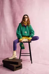 Sudadera con capucha Fruit of the Loom en colaboración con Zara, de color verde con detalles en morado en el interior de la capucha, la cartera de botones y los bordes del bolsillo frontal tipo canguro. Presenta mangas largas, cordones ajustables y un parche con el logo en el centro.