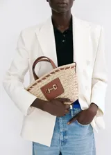 Bolso de mano y bandolera de fibra natural trenzada, color beige, con detalles en cuerina marrón y aplique metálico dorado en el frente. Cuenta con doble asa corta y correa larga desmontable.