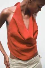 Top de color naranja vibrante con cuello halter que se anuda en la espalda, creando un escote drapeado o fluido en el frente. El bajo del top tiene un corte en pico con tiras finas colgantes.