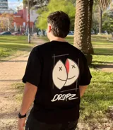 Remera negra de corte relajado con un gran estampado gráfico en la espalda que muestra dos mitades de un rostro sonriente intervenido con cruces en los ojos y un borde rojo. Debajo del gráfico se lee la palabra "DROPZ" en tipografía brush blanca.