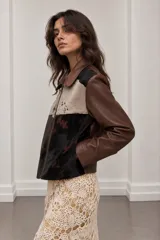 Chaqueta corta de cuero y pelo vacuno, diseño patchwork con bloques en tonos chocolate, negro y crudo, cuello clásico, mangas largas y cierre metálico frontal.