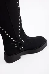 Bota de caña alta confeccionada en cuero vacuno, color negro, con diseño de tachas metálicas dispuestas en forma de arco a lo largo de la caña y correa decorativa con hebilla en la zona del tobillo. Presenta suela track de goma robusta.