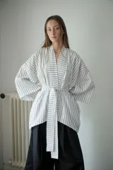 Kimono blanco con rayas verticales negras, confeccionado en tela camisera. Presenta cuello mao, mangas anchas 3/4, bolsillos laterales invisibles y cinturón ajustable desmontable.
