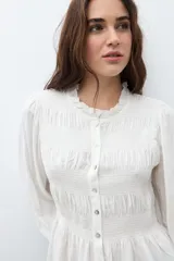 Camisa de color crudo con detalle de fruncido en el pecho, cuello redondo con volado sutil, cierre frontal con botones y mangas largas abullonadas con puños elásticos.