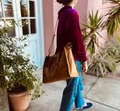 Bolso de mano grande de cuero color marrón, con doble asa corta y correa larga desmontable. Tiene cierre superior y detalles de flecos en el frente.