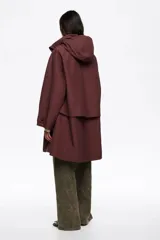 Gabardina midi de corte línea A en color bordó, confeccionada en mezcla de algodón técnico. Presenta cierre frontal con cremallera metálica, cuello subido con capucha integrada y mangas largas ajustables con botones de presión en los puños. Incluye bolsillos laterales y aberturas laterales con botones de presión.