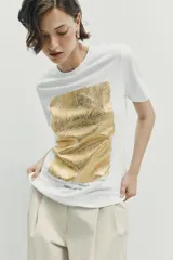 Camiseta de algodón con cuello redondo y manga corta. Presenta un diseño de estampación metalizada en color dorado en la parte frontal con detalles de texto.