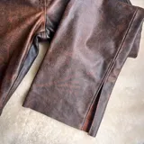 Pantalón de cuero vacuno negro, de corte recto y tiro medio. Presenta costuras horizontales a la altura de la rodilla y aberturas laterales en el ruedo. Incluye un botón de guampa irregular como detalle distintivo en la cintura.