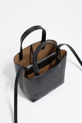 Bolso pequeño de piel vacuna color negro, con estructura rígida, doble asa de mano y correa larga ajustable para llevar al hombro o cruzado. Presenta cierre de botón a presión y logo metálico de la marca en el frente.