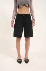 Bermuda de denim negro, tiro bajo, con cintura con trabillas, cierre frontal con cremallera y botón, y bolsillos cargo en el frente.