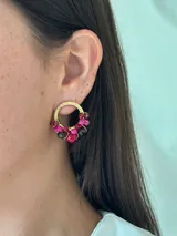 Aro de metal dorado con forma de media luna, del cual cuelgan racimos de cuentas facetadas en tonos fucsia, rosado y granate oscuro.