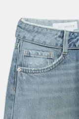 Jeans de mezclilla azul claro, tiro bajo, con corte ancho y pierna recta. Presenta un detalle de cintura cruzada o vuelta con un nudo o pliegue frontal y un ligero desgaste en una de las piernas.