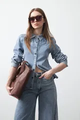 Camisa de jean color azul claro con tachas metálicas en el canesú y a lo largo de las costuras frontales. Tiene mangas largas con puños abullonados y cierre frontal con botones.