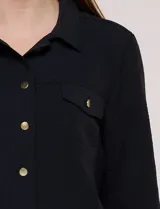 Camperita liviana de corte recto y acabado crepé color blanco, con cuello camisero clásico y cierre frontal de botones metálicos dorados. Presenta bolsillos en el pecho con solapa y mangas largas con puños abotonados.