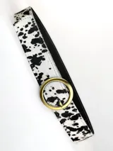 Cinturón de cuero genuino con estampado de manchas tipo vaca en blanco y negro, con hebilla circular metálica en tono bronce.