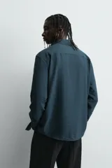 Camisa de corte relajado confeccionada en tejido fluido con mezcla de viscosa. Presenta cuello italiano, manga larga con puños abotonados y cierre frontal con botonadura, destacando por el detalle de jaretas verticales a ambos lados de la tapeta.