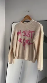 Sweater de punto color beige con diseño de texto en color rosa que dice 'I'm just a girl' y una estrella. Presenta cuello redondo, manga larga y corte holgado.