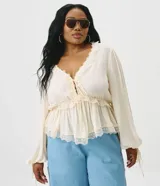 Blusa de viscosa texturada color marfil, con escote en V pronunciado, lazo frontal y corte estilo túnica. Presenta mangas largas abullonadas con puños ajustables mediante tiras y detalles de encaje en el escote, la cintura fruncida y el ruedo.