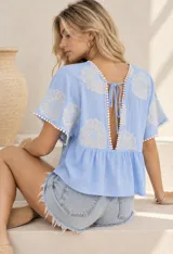 Blusa blanca de corte amplio y corto, con escote en V y mangas cortas. Presenta detalles de bordados florales en los hombros y mangas, además de ribetes con terminación de pompones pequeños.