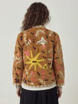 Chaqueta corta de terciopelo color camel con bordados coloridos de inspiración folclórica que incluyen pájaros blancos, corazones rosados, estrellas amarillas y figuras, con cuello redondo y diseño abierto al frente.