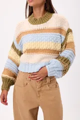 Buzo de tejido de punto grueso con diseño de rayas horizontales multicolores en tonos blanco, rosa, verde oliva y beige. Presenta cuello redondo acanalado, mangas largas con puños ajustados y un corte holgado.