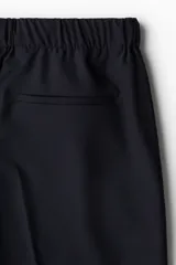 Pantalón negro de corte relajado, confeccionado en tejido suave. Presenta cintura elástica con cordón oculto, cierre con botón y corchete, bolsillos laterales y bolsillos insertados traseros. Las piernas son ajustadas y tienen pliegues marcados.