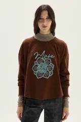 Buzo oversize de felpa color marrón con bordado frontal de una flor estilizada y la palabra "Muse" escrita en cursiva en color celeste.
