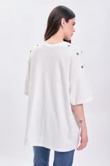 Remera blanca de corte holgado con cuello redondo y mangas cortas. Presenta detalle de ojales metálicos en los hombros.