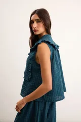 Blusa de algodón con patrón a cuadros vichy en tonos verde oscuro y azul, sin mangas, con cuello amplio decorado con volados, botones delanteros y un bolsillo frontal aplicado con ribete de volados.