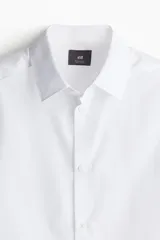 Camisa blanca de manga larga con cuello inglés, tapeta lisa y bajo redondeado. Presenta canesú en la espalda y puños ajustables con botón.