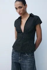 Camisa entallada de popelín negro con estampado de lunares blancos, escote en pico, cuello solapa y mangas muy cortas. Cuenta con costuras marcadas y cierre lateral con cremallera oculta.