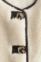 Chaleco de efecto peludito color beige, con cuello redondo, cierre frontal de botones y ribetes en contraste marrón oscuro. Incluye bolsillos laterales discretos y forro interior.