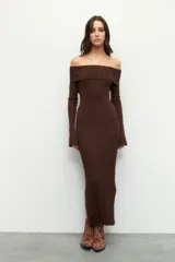 Vestido largo de punto acanalado en color negro, con diseño de hombros descubiertos y mangas largas. Presenta un corte ajustado al cuerpo y aberturas laterales en el ruedo.