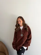 Buzo de corderito color marrón, con cuello alto y cierre frontal parcial. Presenta un diseño holgado con mangas largas y un parche decorativo en el frente.